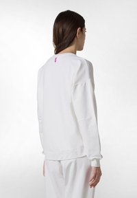 Vit sweatshirt gjord av mjukt tyg med avslappnad passform, som har neddragna axlar och en liten rosa etikett på baksidan.