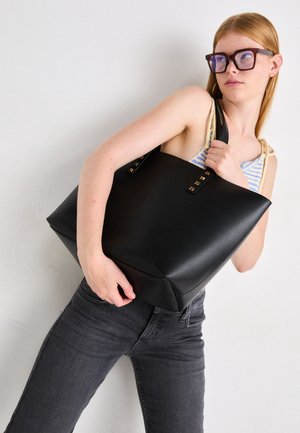 Junge Frau mit Brille hält eine große schwarze Einkaufstasche über der Schulter, trägt ein gestreiftes Oberteil und dunkle Jeans vor neutralem Hintergrund.