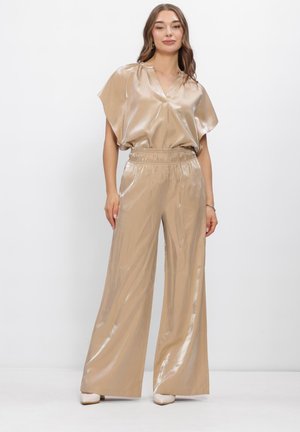 Vrouw staand in beige glanzende wijde broek en bijpassende blouse met korte mouwen en elastische taille op een witte achtergrond.