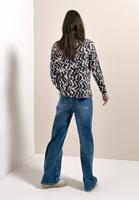 Mujer de pie frente a una pared beige, vistiendo una blusa de manga larga con estampado de zigzag en blanco y negro, vaqueros anchos azules y zapatos beige.