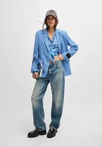 Helles blaues, strukturiertes Blazer über einem blauen Batik-Top, kombiniert mit ausgewaschenen Jeans und klobigen schwarzen Schuhen, ergänzt durch eine graue Mütze.