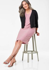 Rosa, knielanges Kleid mit einer taillierten Form, kombiniert mit einem schwarzen, strukturierten Cardigan. Silberne, verzierte High Heels runden den Look ab.