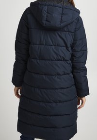 Femme portant un long manteau en duvet bleu marine matelassé avec capuche, vue de dos sur un fond uni.