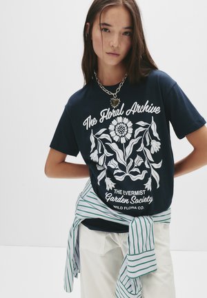 Jeune femme portant un t-shirt marine avec un imprimé floral blanc portant la mention "L'Archive Florale" et une chemise rayée nouée autour de la taille.