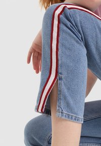 Jeans de mezclilla azul con bandas laterales rojas y blancas, con un diseño recortado y detalles de dobladillo dividido en los puños. Textura suave.