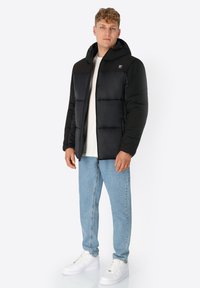 Schwarze Puffjacke mit Kapuze, ausgestattet mit einem Reißverschluss und gestepptem Design. Getragen über einem weißen Hemd und kombiniert mit hellblauen Jeans und weißen Turnschuhen.