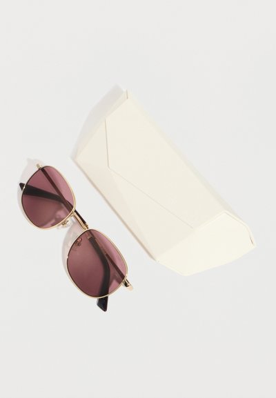 Γυαλιά ηλίου με χρυσαφένιο περίγραμμα και φακούς με μοβ απόχρωση, τοποθετημένα δίπλα σε λευκή γωνιακή θήκη γυαλιών σε ανοιχτό φόντο.