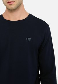 Maglietta a maniche lunghe navy in tessuto morbido con un piccolo logo grigio sul petto, collo rotondo e texture liscia.