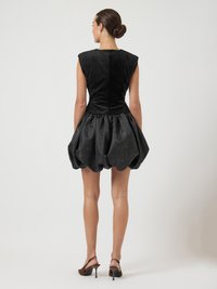 Mulher com cabelo preso em um coque, usando um vestido preto sem mangas com corpete justo e saia texturizada volumosa, de pé contra um fundo branco liso.