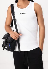 White ribbed tank top with a round neckline, featuring a black "KARL LAGERFELD" logo. Worn with black sweatpants and a black satchel bag.