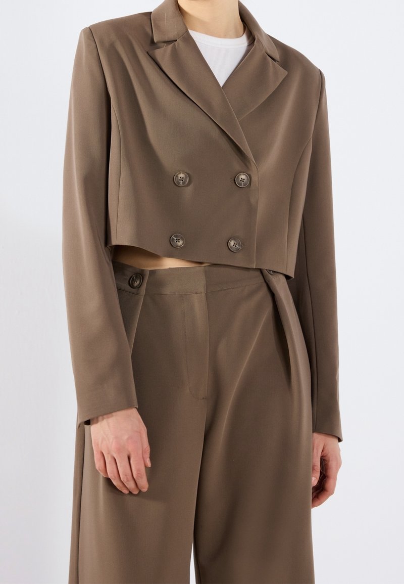 Brązowa krótka marynarka dwurzędowa z wyciętym kołnierzem i dwoma guzikami. Dopasowane spodnie z wysokim stanem o gładkiej fakturze.