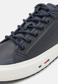 Zbliżenie granatowego buta sportowego ze skóry z dopasowanymi sznurowadłami, białą podeszwą z małymi czerwonymi, białymi i niebieskimi detalami oraz wytłoczonym logo po boku.