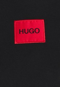 Tela negra con un tejido texturizado que presenta una etiqueta rectangular roja prominente con la palabra "HUGO" en bordado negro.