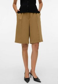 Lichtbruine plooishorts met een losse pasvorm en zijzakken, gecombineerd met een zwarte, knopen-down blouse en zwarte puntige hakken.