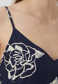 Námořnická modrá látka s bílým květinovým vzorem má design s výstřihem do V a tenkými ramínky, což zdůrazňuje hladkou strukturu a jednoduché detaily.