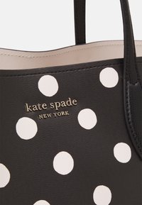 Borsa nera con grandi pois bianchi, caratterizzata da un logo dorato che reca la scritta "kate spade NEW YORK", realizzata in materiale testurizzato e dotata di due manici.
