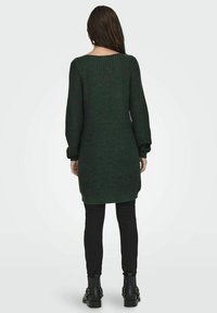 Lungo maglione lavorato a maglia verde scuro con colletto a costine e maniche a palloncino, indossato sopra pantaloni neri aderenti e stivaletti. Tessuto texture.