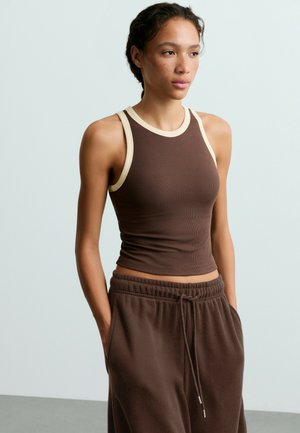 Une femme porte un débardeur côtelé marron bordé de crème et un pantalon assorti à cordon de serrage, devant un fond de studio clair.