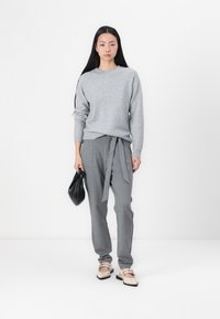 Grauer Strickpullover mit langen Ärmeln, kombiniert mit grauen, mit Gürtel versehenen Hosen. Das Modell hält eine schwarze Tasche und trägt helle flache Schuhe.
