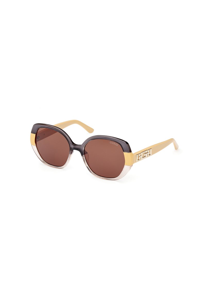 Gafas de sol con lentes octagonales de gran tamaño. La montura es de color marrón oscuro con un borde inferior transparente y brazos amarillos, con un acento de logo en un lado.