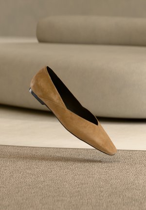 Tan suède platte schoen met spits toelopende neus en zwarte zool, zwevend boven een gestructureerd beige tapijt tegen een neutrale bank als achtergrond.