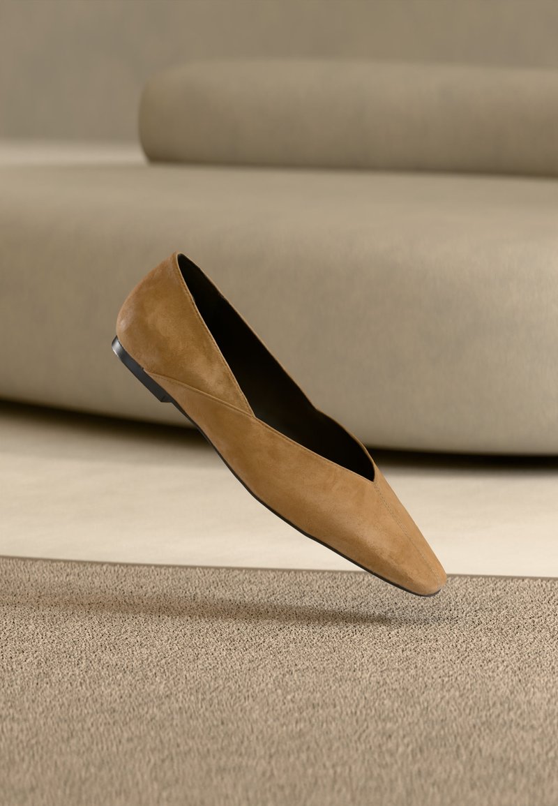 Chaussure plate pointue en daim couleur chameau avec semelle noire, flottant au-dessus d'un tapis beige texturé devant un fond de canapé neutre.