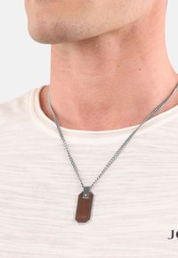 Collana in argento con un ciondolo rettangolare caratterizzato da un'intarsio in legno e accenti in argento, indossata sopra una maglietta leggera e testurizzata.