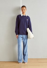 Μπλούζα navy με μικρό κεντημένο λογότυπο, σε συνδυασμό με ανοιχτό μπλε παν�τελόνι σε φαρδιά γραμμή και λευκή τσάντα φάκελο. Ξύλινο δάπεδο και απλό φόντο.