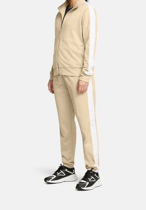 Person iført beige træningsdragt med hvide striber på ærmer og bukser samt sorte og hvide sportssko, stående i profil.