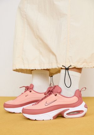 Sneakers Nike in tonalità di rosa e pesca con un design texturizzato, suole bianche e ammortizzazione ad aria visibile. Indossati con calzini bianchi a metà polpaccio.