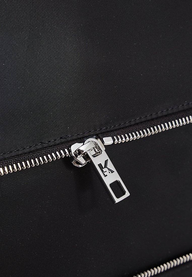 Bolso de tela negra con detalles de cremallera plateados. Presenta un tirador de cremallera prominente con el logo y costuras de contraste a lo largo de las costuras.