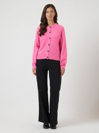 Mulher de pé, vestindo um cardigã rosa choque abotoado, calças largas pretas e sapatos pretos, contra um fundo claro liso.