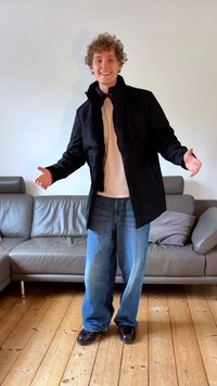 Μαύρο μπουφάν με ψηλό γιακά, φορεμένο πάνω από ανοιχτόχρωμο πουκάμισο. Χαλαρά μπλε τζιν και καφέ παπούτσια. Ξυλοτύπος δαπέδου, σύγχρονος γκρι καναπές στο background.