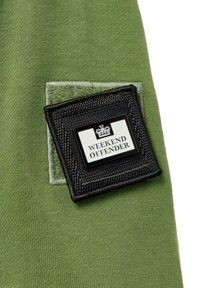 Green fabric with a smooth texture features a black patch with a white logo reading "WEEKEND OFFENDER," bordered by a soft green felt detail.
