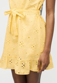 Κίτρινο φόρεμα με οπές, κεντήματα με λουλούδια, βολάν στο τελείωμα και δεμένη ζώνη στη μέση στο κάτω μέρος του κορμού και το χέρι μιας γυναίκας.