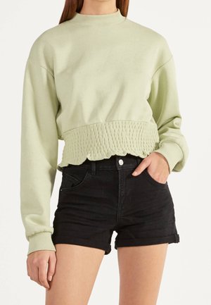 Frau trägt ein hellgrünes, langärmeliges Cropped-Sweatshirt mit strukturiertem Saum und schwarze, hoch taillierte Jeans-Shorts mit aufgekrempeltem Saum.