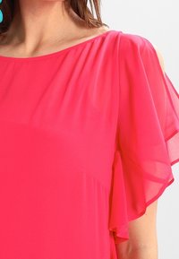 Haut en chiffon rose vif avec un design épaules dénudées, des manches volantées, une texture lisse et un drapé doux. Coupe ajustée et fluide sur le corps.