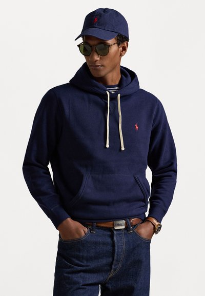 Sudadera de algodón azul marino con un bolsillo canguro, cordones de contraste en color crema y un pequeño logo rojo. Combinada con jeans oscuros.