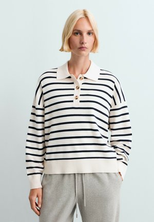 Femme blonde portant un pull polo à manches longues rayé blanc et bleu marine et un pantalon gris clair avec cordon, debout devant un fond uni.