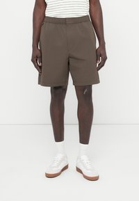 Καφέ σορτς με ελαστική μέση και κούμπωμα με κουμπί, με πλαϊνές τσέπες και άνετη εφαρμογή, συνδυασμένα με λευκά αθλητικά παπούτσια και κάλτσες.