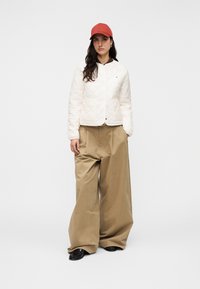 Λευκό μπουφάν με καπιτονέ ύφασμα και στρογγυλή λαιμόκοψη, φαρδιά καφέ παντελόνια και κόκκινο καπέλο. Το μπουφάν διαθέτει εμπρόσθιο κλείσιμο με κλιπ και διακριτική λεπτομέρεια με λογότυπο.