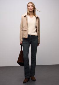 Chaqueta beige de mezcla de lana con cuello, suéter de pico color crema, vaqueros acampanados gris oscuro y botines marrones, sosteniendo un bolso de ante marrón.
