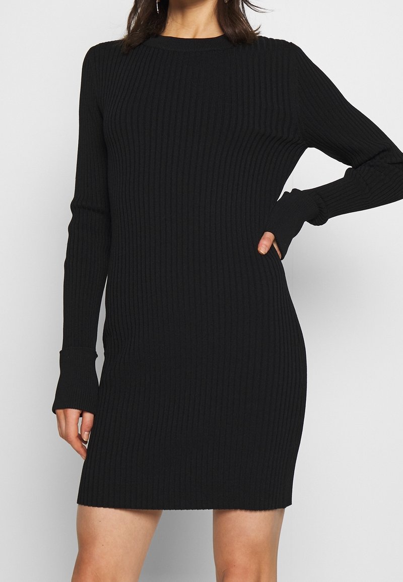 Femme portant une robe en tricot côtelé noire à manches longues avec un col rond et la main sur la hanche, debout devant un fond uni.