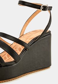 Sandale compensée noire avec trois brides sur le devant, une bride de cheville avec boucle en argent, et semelle intérieure en motif beige sur fond blanc.