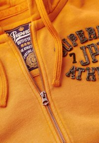 Felpa arancione con zip realizzata in tessuto morbido. Presenta lettere testurizzate, una zip in metallo e un'etichetta interna con marchio.
