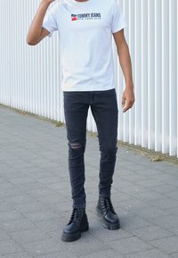 Vit bomulls-t-shirt med "TOMMY JEANS"-logotyp, i kombination med slitna svarta skinny jeans och svarta snörda platåstövlar. Slät textur.