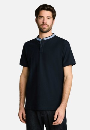 Homme portant un t-shirt henley à manches courtes bleu marine foncé avec un col bleu clair et un pantalon foncé, debout devant un fond uni.