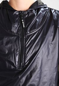 Plano cercano de una persona que lleva una chaqueta negra brillante con cremallera frontal etiquetada "G-STAR" y una capucha verde oscura debajo.