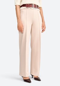 Pantaloni a gamba larga di un rosa chiaro, realizzati in tessuto liscio, con dettagli di piega frontale. Abbinati a una cintura marrone scuro ornata con borchie argentate.