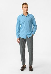 Lichtblauwe overhemd met lange mouwen en kraag, gecombineerd met grijze op maat gemaakte broek en bruine loafers, tegen een neutrale achtergrond.